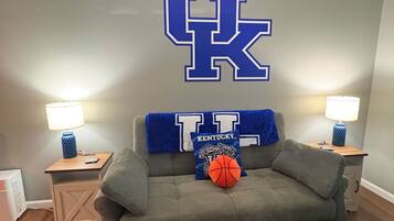 Ein Zimmer mit einem grauen Sofa, zwei Nachttischen mit Lampen, einer großen Wanddekoration mit den Buchstaben 'UK' und einem Basketball auf dem Sofa.