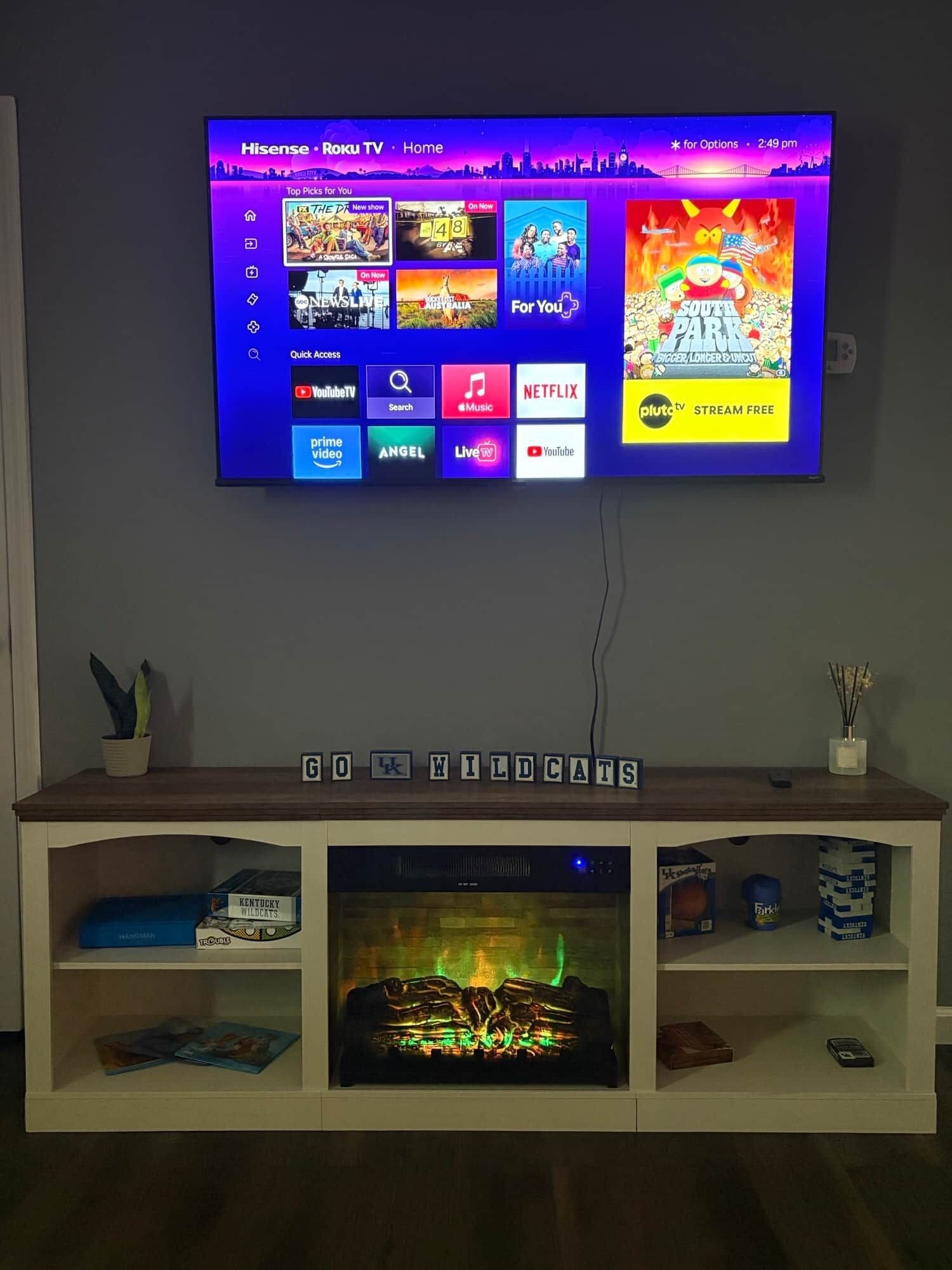 Un téléviseur fixé au mur affiche l’interface Roku TV, avec divers logos de services de diffusion en continu. Sous le téléviseur, une étagère présente des jeux de société et un objet décoratif.