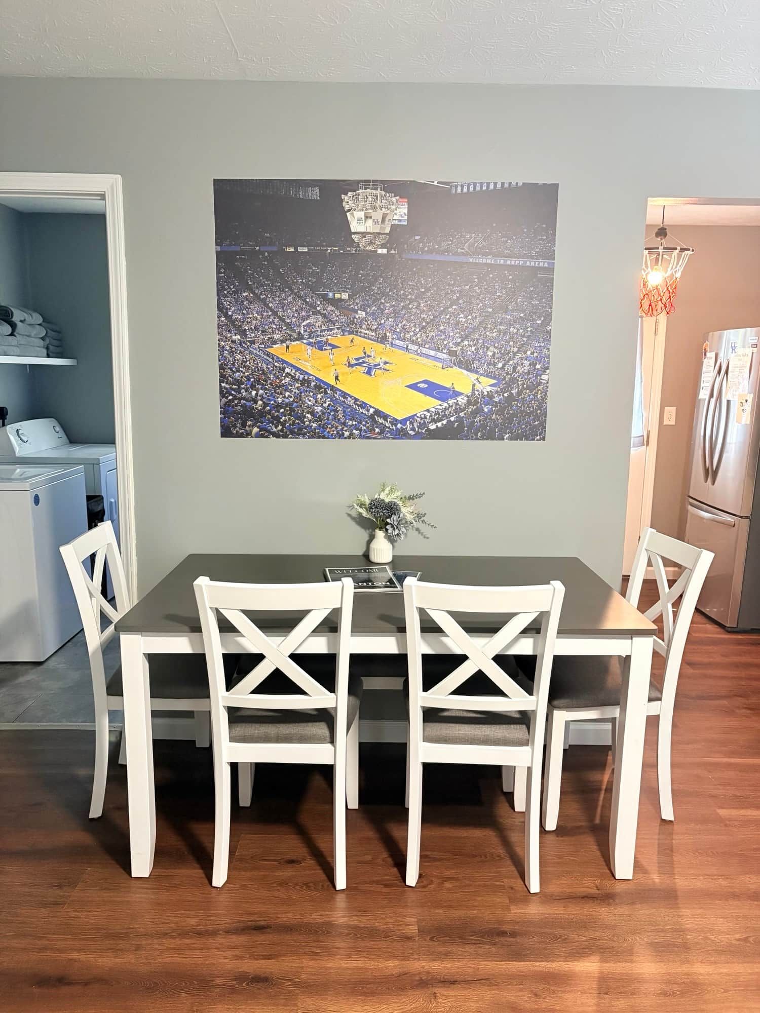 Un coin repas avec une table et des chaises, une affiche sportive accrochée au mur, une petite plante et un espace cuisine équipé d’appareils électroménagers.