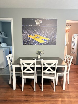 Un coin repas avec une table et des chaises, une affiche sportive accrochée au mur, une petite plante et un espace cuisine équipé d’appareils électroménagers.