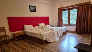 Un dormitorio con piso de madera, una cama con ropa de cama blanca, cabecera roja, mesita de noche, una silla y una ventana con cortinas.