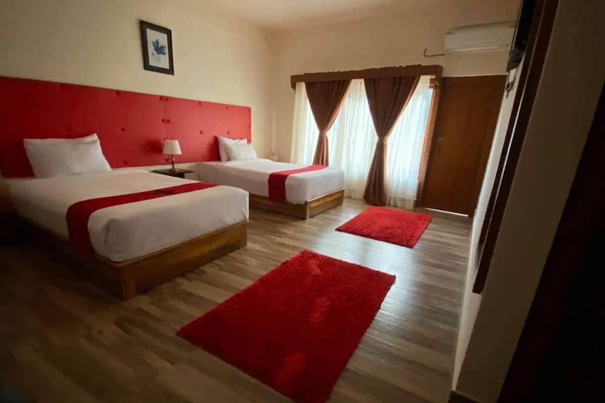 A hotel room with two beds, a red headboard, a wooden floor, and a window with curtains.