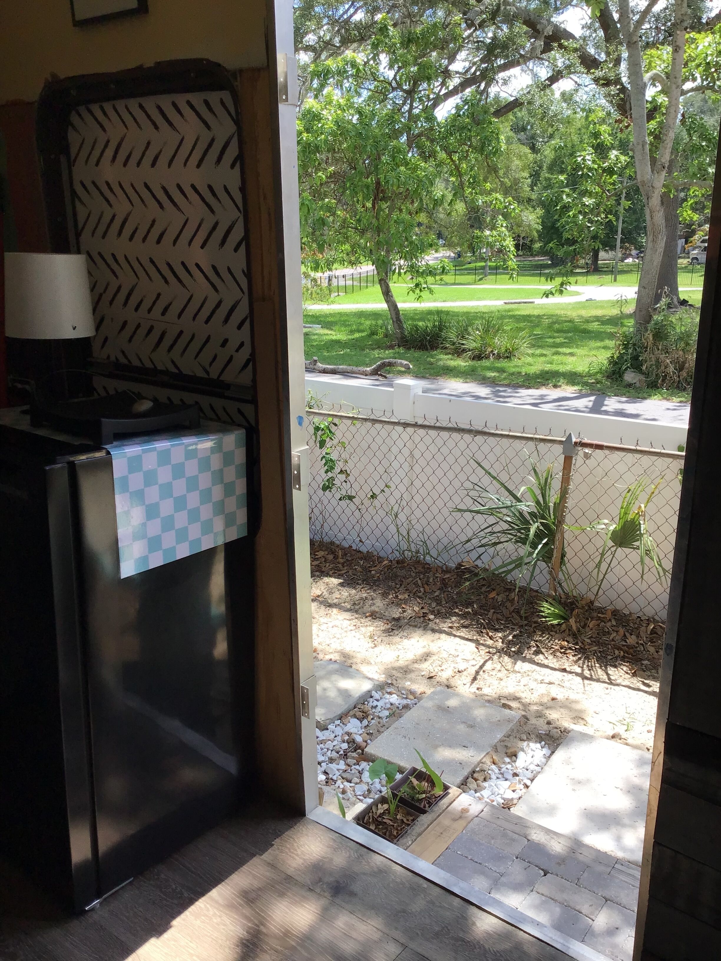 An open door to a room with a kitchen counter, a checkered tablecloth, a lamp, and a view of a grassy area with trees and a fence.