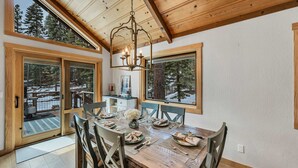 A dining area with a wooden table set for four, large windows, and a chandelier. - Tahoe City Escape | Beach Access + Large Deck (Tahoe City)