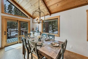 A dining area with a wooden table set for four, large windows, and a chandelier. - Tahoe City Escape | Beach Access + Large Deck (Tahoe City)