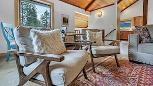 A spacious living area with wooden ceiling beams, a large window with a view of trees, and a cozy seating arrangement with plush cushions. - Tahoe City Escape | Beach Access + Large Deck (Tahoe City)