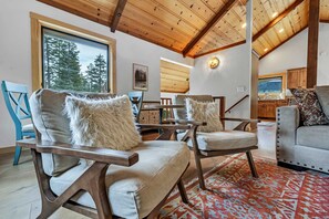 A spacious living area with wooden ceiling beams, a large window with a view of trees, and a cozy seating arrangement with plush cushions. - Tahoe City Escape | Beach Access + Large Deck (Tahoe City)