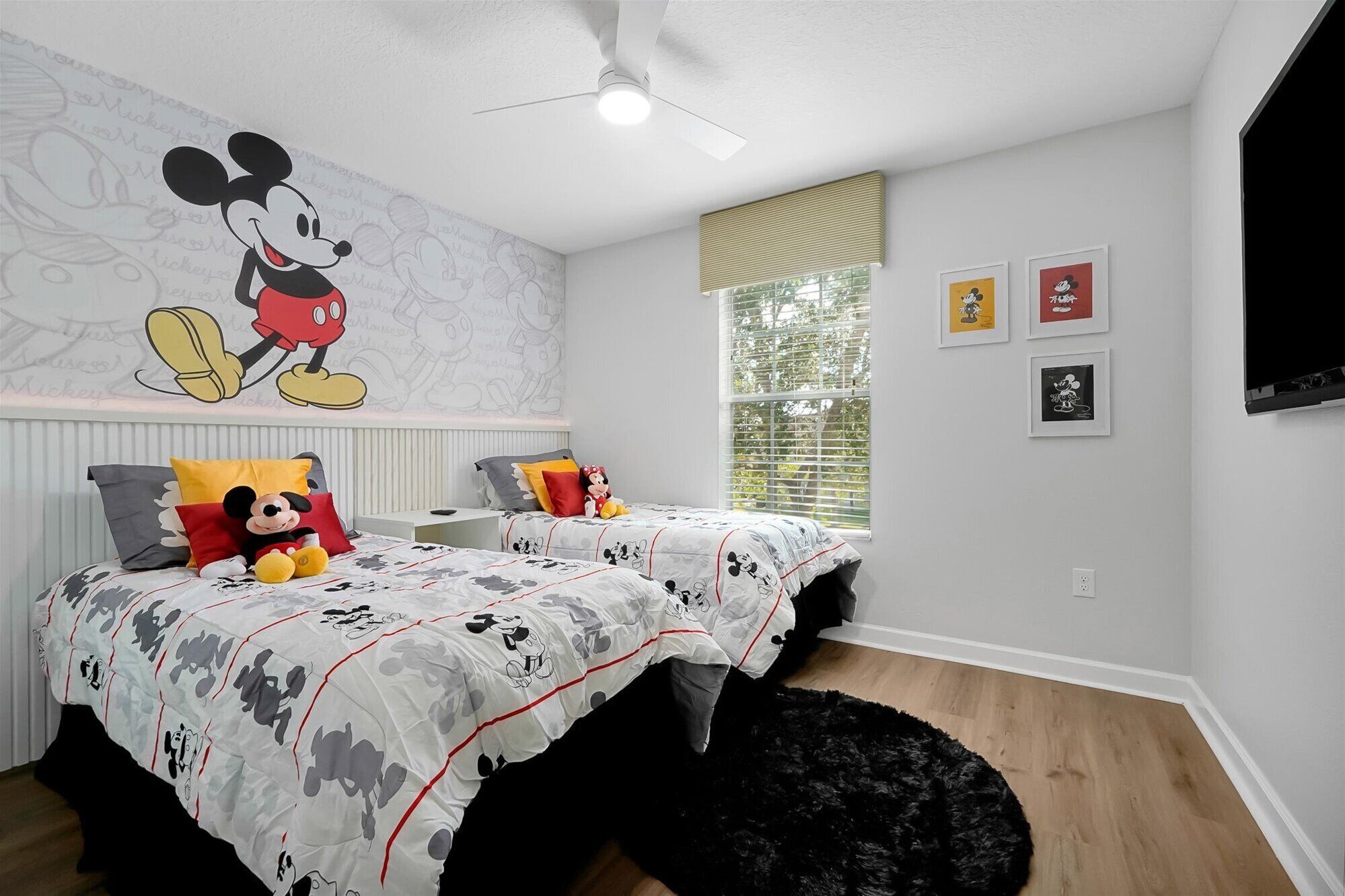 A bedroom with Mickey Mouse wallpaper, a bed with Mickey-themed bedding, a black furry rug, a window with blinds, and framed pictures on the wall.