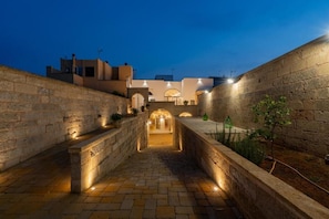 Une cour bordée de murs en pierre, avec un chemin central menant à l’entrée d’un bâtiment, le tout éclairé par une lumière chaleureuse.