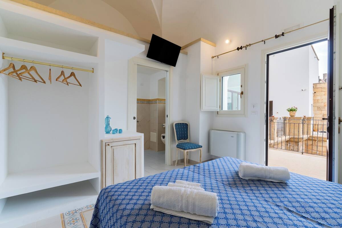 Ein Schlafzimmer mit Bett, Sessel, an der Wand montiertem Fernseher und Balkon, der durch eine offene Tür sichtbar ist.