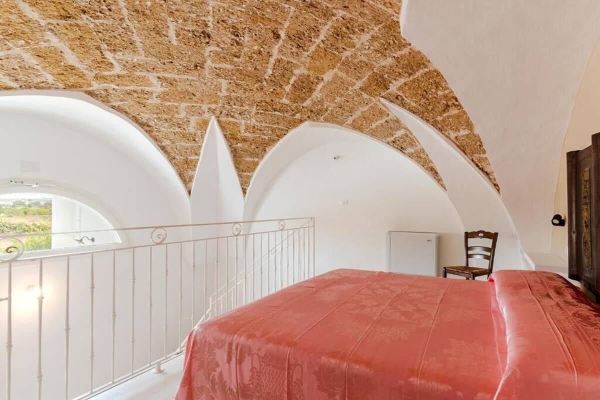 Ein Schlafzimmer mit rotem Bettbezug, weißen Wänden und einer Ziegeldecke. Es gibt einen Balkon mit weißer Brüstung und einem Stuhl.