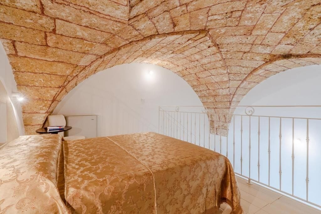 A bedroom with a vaulted stone ceiling, a bed with a patterned bedspread, a small bedside table, and a view of the outside through a balcony railing.