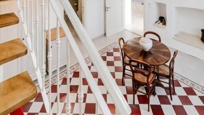 Ein Zimmer mit kariertem Boden, einer Treppe mit weißen Geländern, einem runden Holztisch mit Stühlen und einem Bett mit rosa Decke.