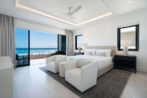 Une chambre d’hôtel moderne avec un grand lit, deux fauteuils, une table de chevet, un ventilateur de plafond et une vue sur l’océan.
