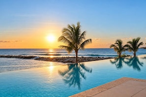 A clear view of a swimming pool with a sunset in the background, palm trees, and a rocky shoreline. - Ocean Front Staffed Villa | Lux Resort by Bocobay (St. Ann’s Bay)