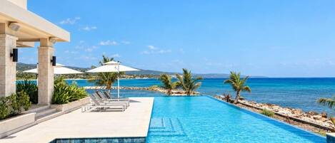 A swimming pool with a view of the ocean, lounge chairs, and palm trees.