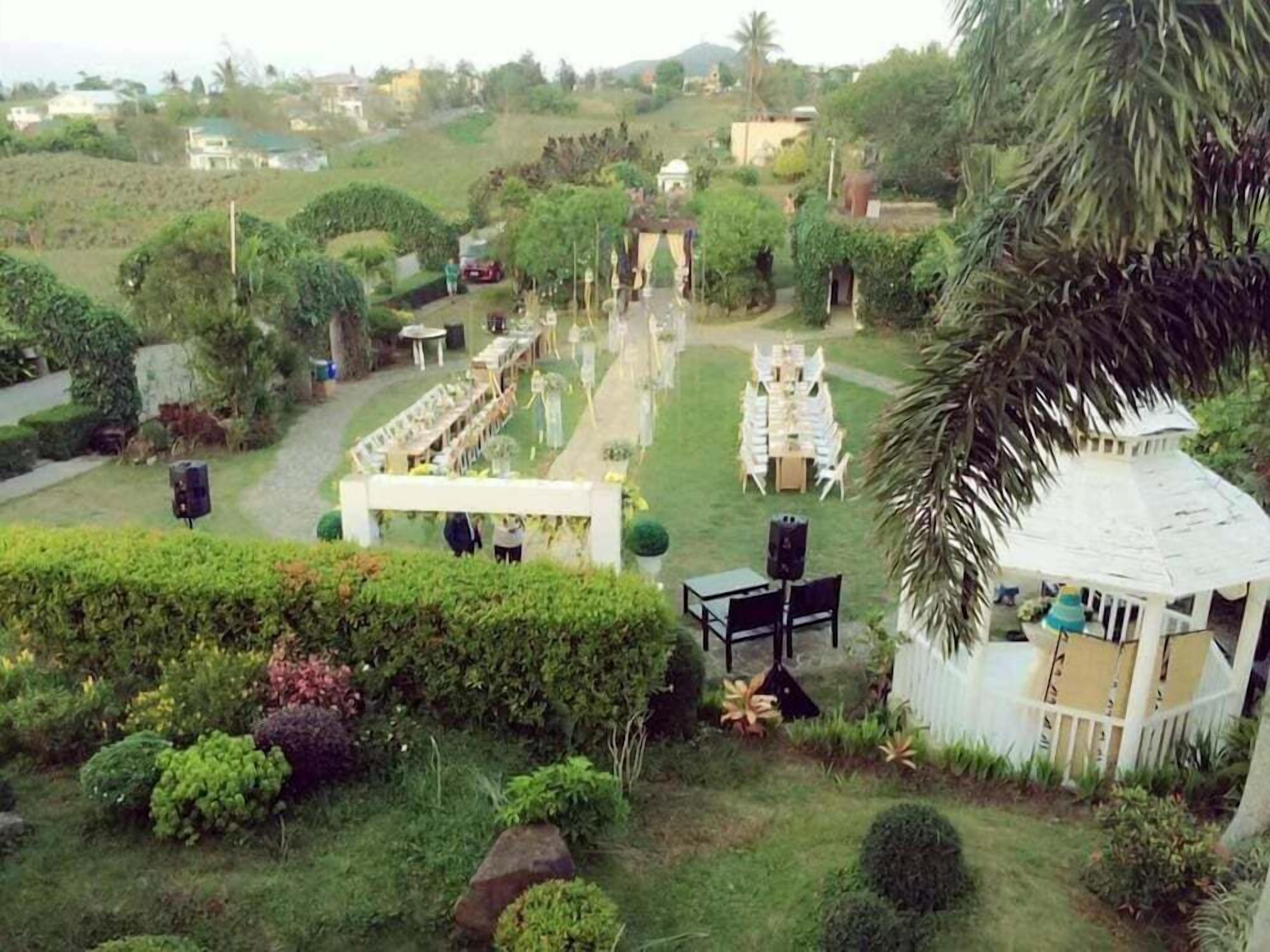 Un jardín con un pabellón blanco, una zona de comedor con mesas y sillas, y un sendero que conduce a un edificio blanco.