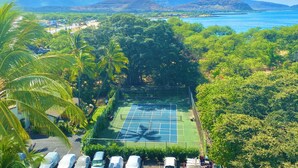 A tennis court surrounded by lush greenery and palm trees, with mountains and a body of water in the background. - Ocean-Front Condo•Semi-Private Beach•Pool•Hot Tub•Tennis.Free Parking. (Waianae)