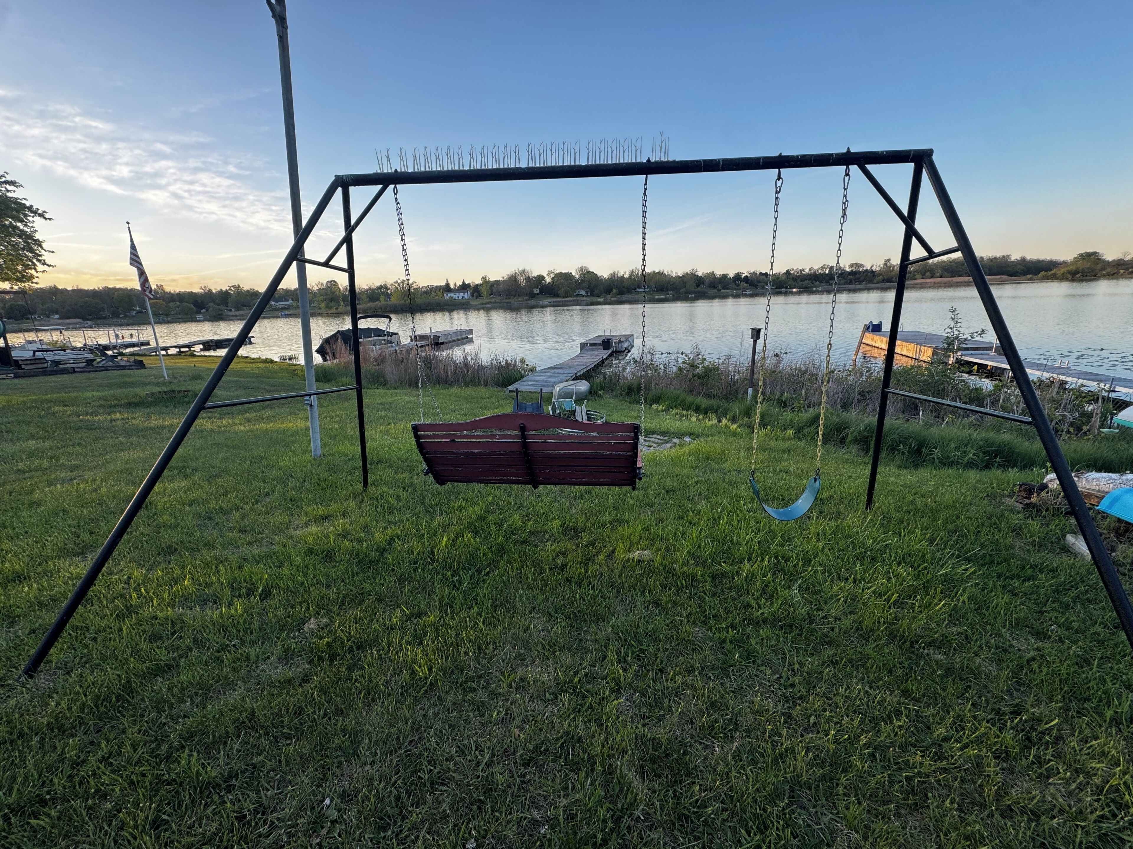 Spielplatz mit Schaukeln und Bank in einer Wiese am See, im Hintergrund liegen Boote.