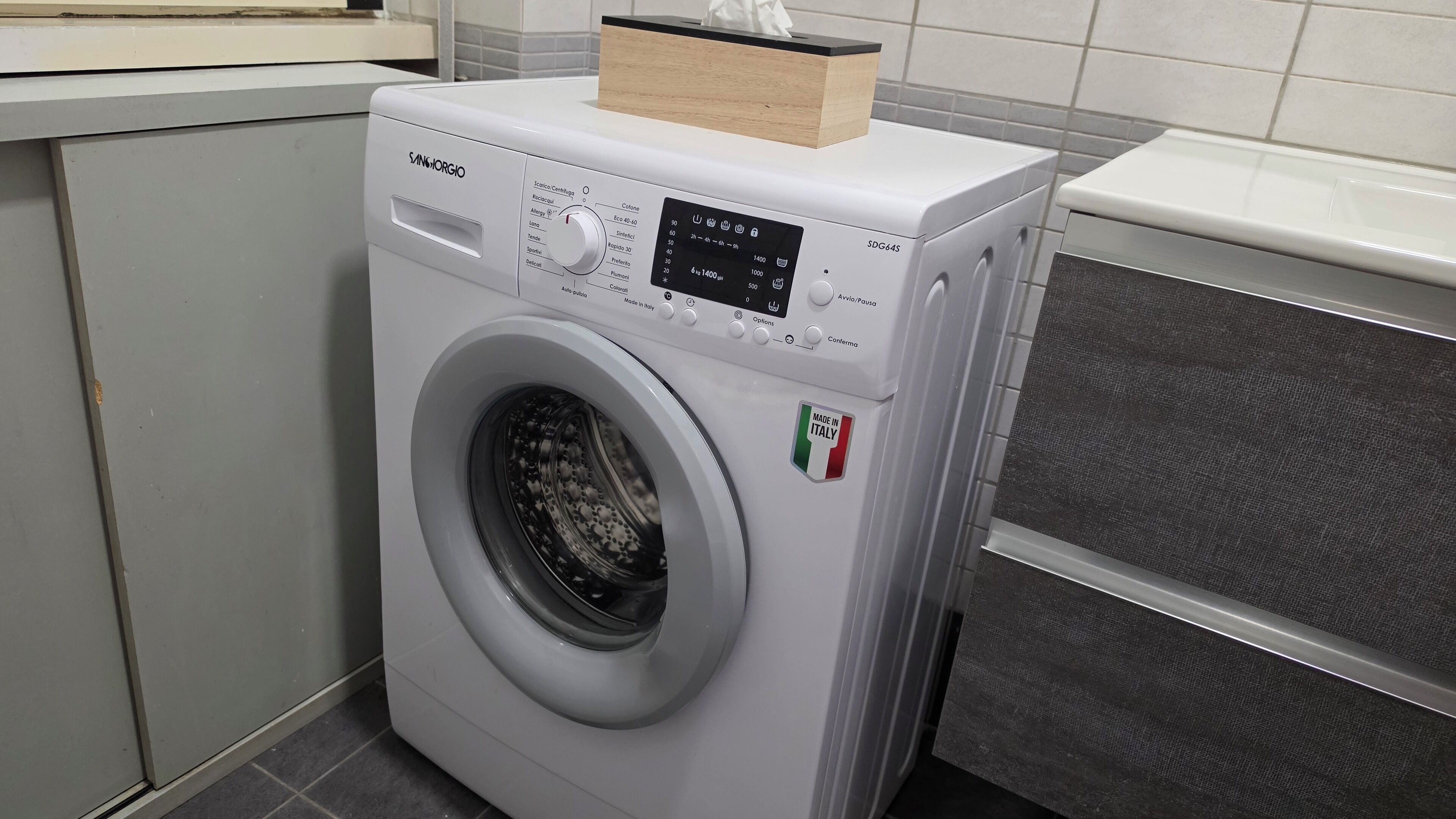 Une machine à laver blanche, dotée d’un panneau de commande et surmontée d’une boîte en bois, avec une étiquette indiquant « Fabriqué en Italie ».