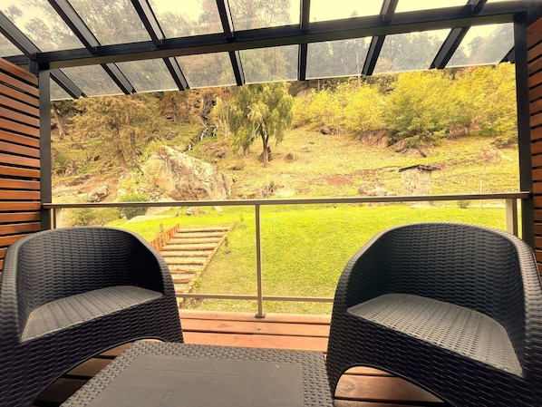 A covered outdoor seating area with two wicker chairs and a table, overlooking a natural landscape with a grassy area and trees.