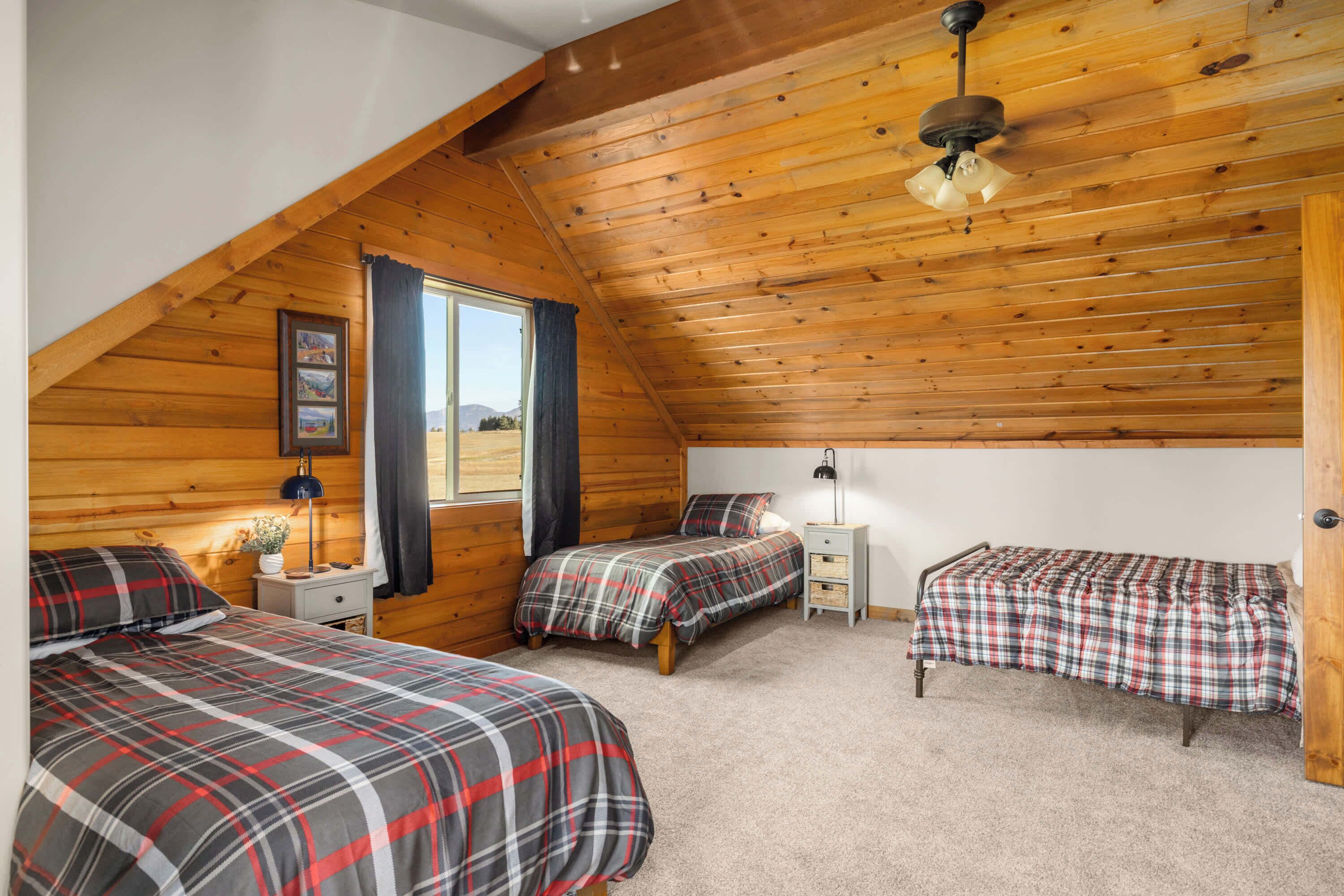 A cozy attic bedroom with wooden walls, a sloped ceiling, and three beds with plaid bedding.