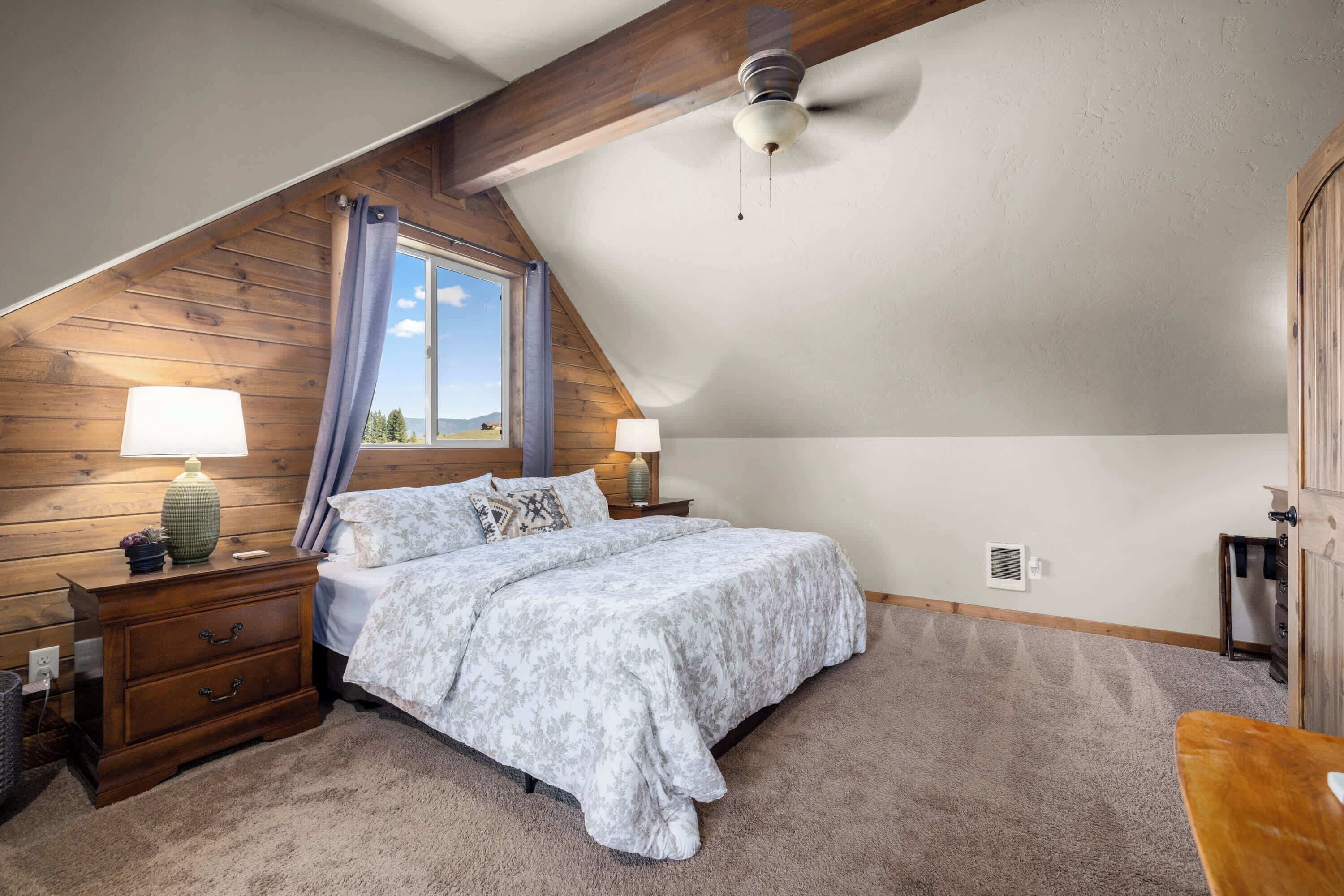 A bedroom with a sloped ceiling, a bed with a patterned bedspread, two bedside lamps, a wooden nightstand, and a window with curtains.