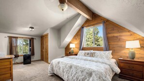 A bedroom with a sloped ceiling, a bed with a floral bedspread, two bedside lamps, a wooden dresser, and a window with curtains. - Glacier River Ranch - Flathead (Columbia Falls)