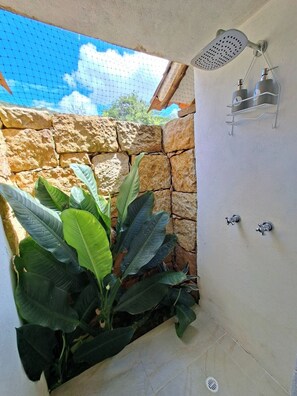 Un espace douche avec un mur en pierre, une pomme de douche suspendue et une Ă©tagĂšre en mĂ©tal oĂč sont posĂ©es des bouteilles.