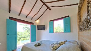 A bedroom with a bed, two windows with shutters, a TV, and a view of greenery outside.