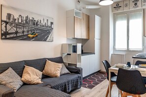 A modern living room with a sofa, a dining table with chairs, a kitchen area, and a cityscape painting on the wall. - [Savona Old Town] Cruises 10 mins • Free WIFI (Savona)