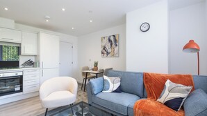 Ein modernes Wohnzimmer mit Sofa, Sessel und Couchtisch. Es gibt eine Küchenzeile mit Schränken und einem Esstisch.