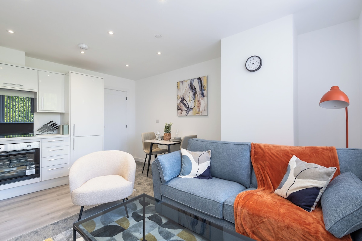Una sala de estar moderna con sofá, sillón y mesa de centro. Se observa una zona de cocina con armarios y una mesa de comedor.