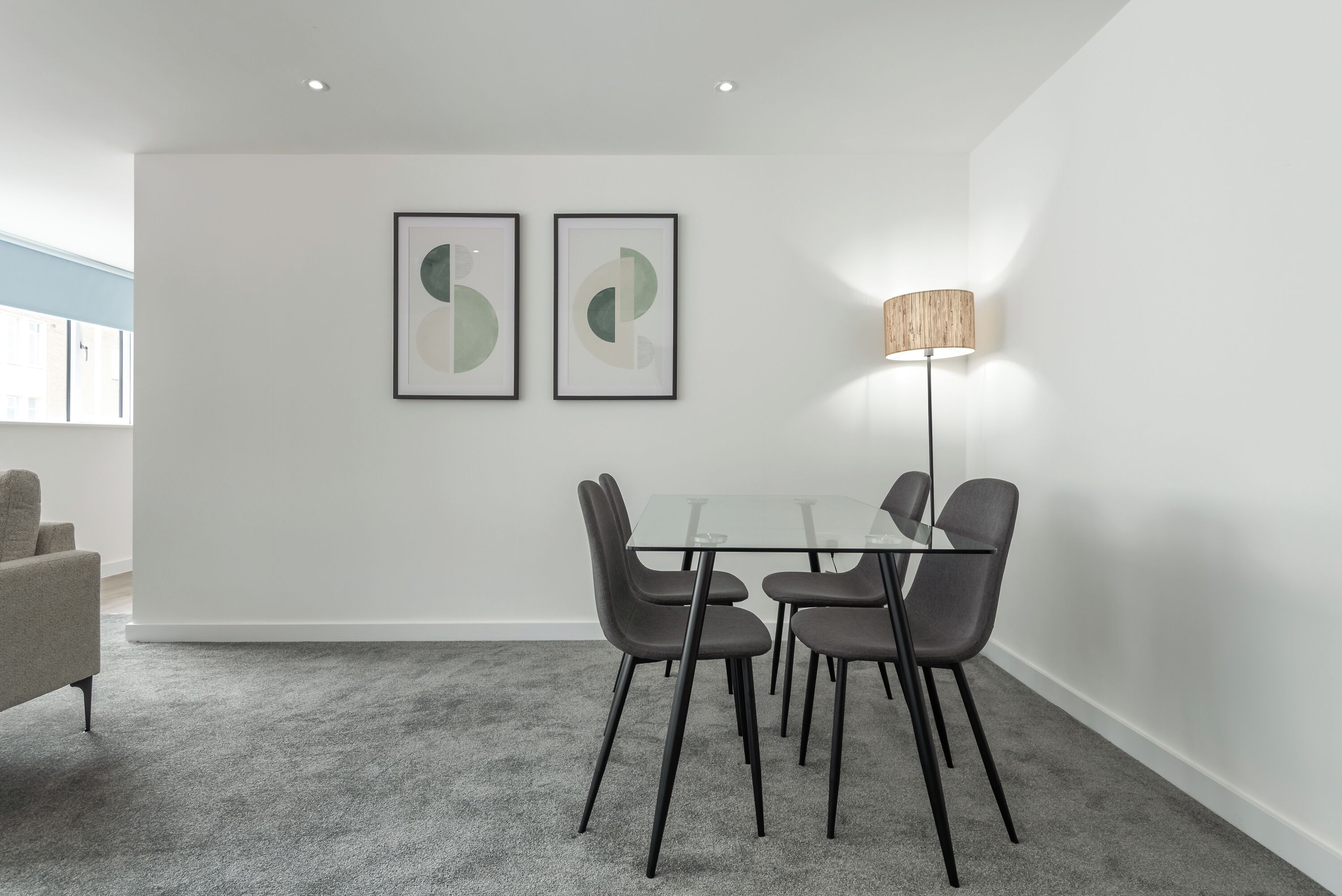 Un comedor moderno con una mesa de vidrio y cuatro sillas, una lámpara de pie y dos obras de arte abstracto en la pared.