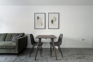 Une pièce avec un canapé vert, deux œuvres d’art abstraites encadrées, une table en bois avec quatre chaises et un sol recouvert d’une moquette.