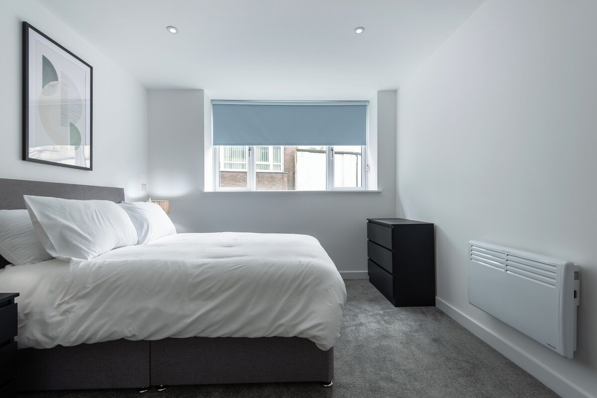 Un dormitorio moderno con cama, mesita de noche, cómoda, ventana con persianas y radiador.
