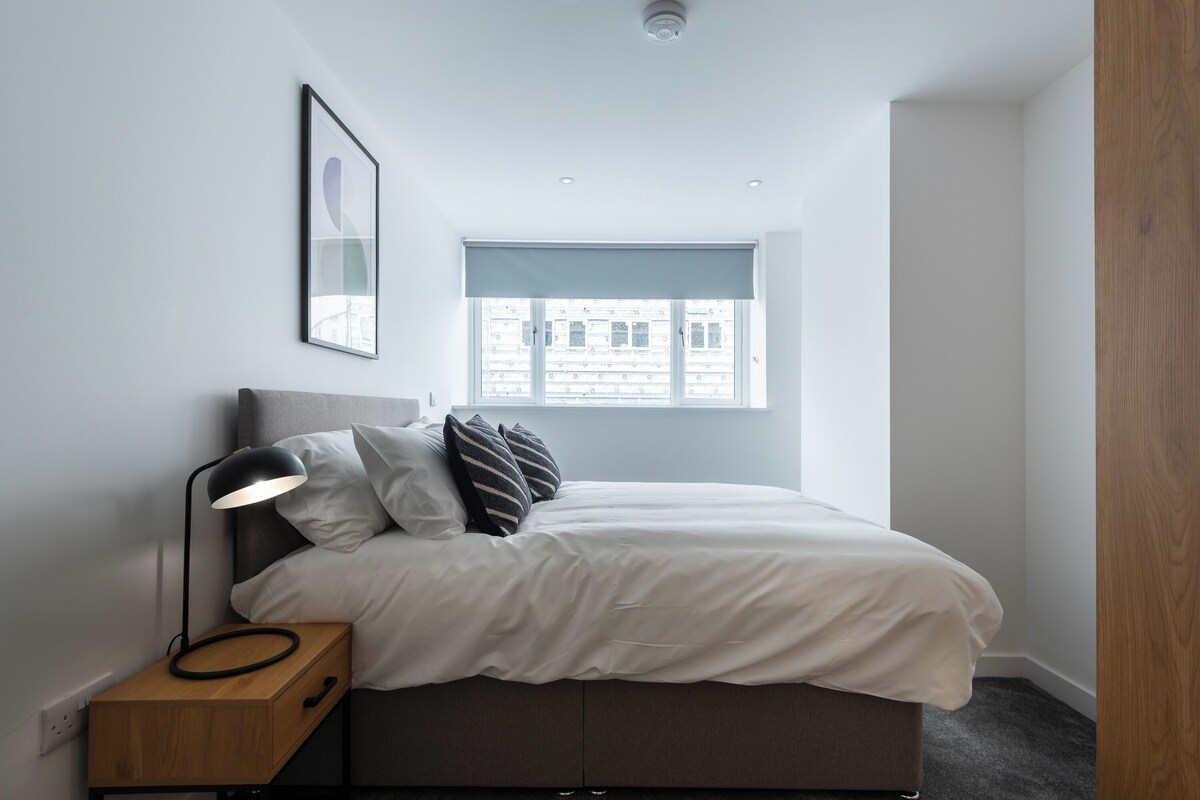 Un dormitorio con cama, mesita de noche, lámpara, ventana y un cuadro enmarcado en la pared.