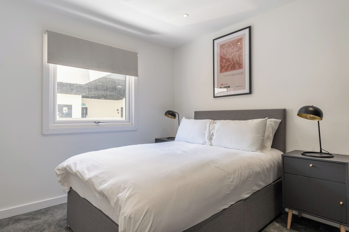 Un dormitorio con cama, mesitas de noche, una ventana y un cuadro en la pared.