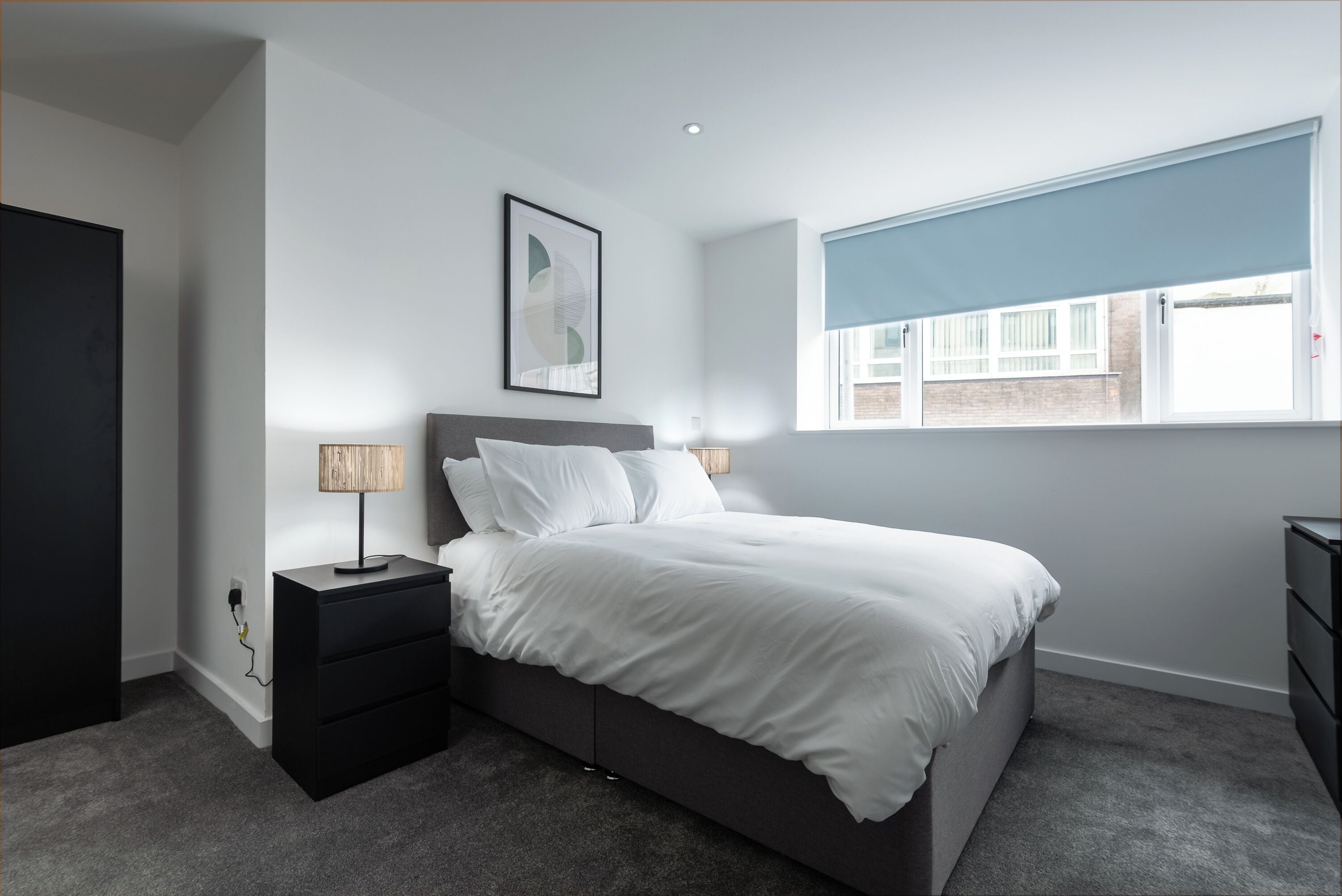 A modern bedroom with a bed, bedside tables, a lamp, a window with blinds, and a wall-mounted artwork.