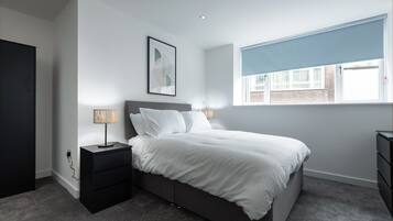 A modern bedroom with a bed, bedside tables, a lamp, a window with blinds, and a wall-mounted artwork.