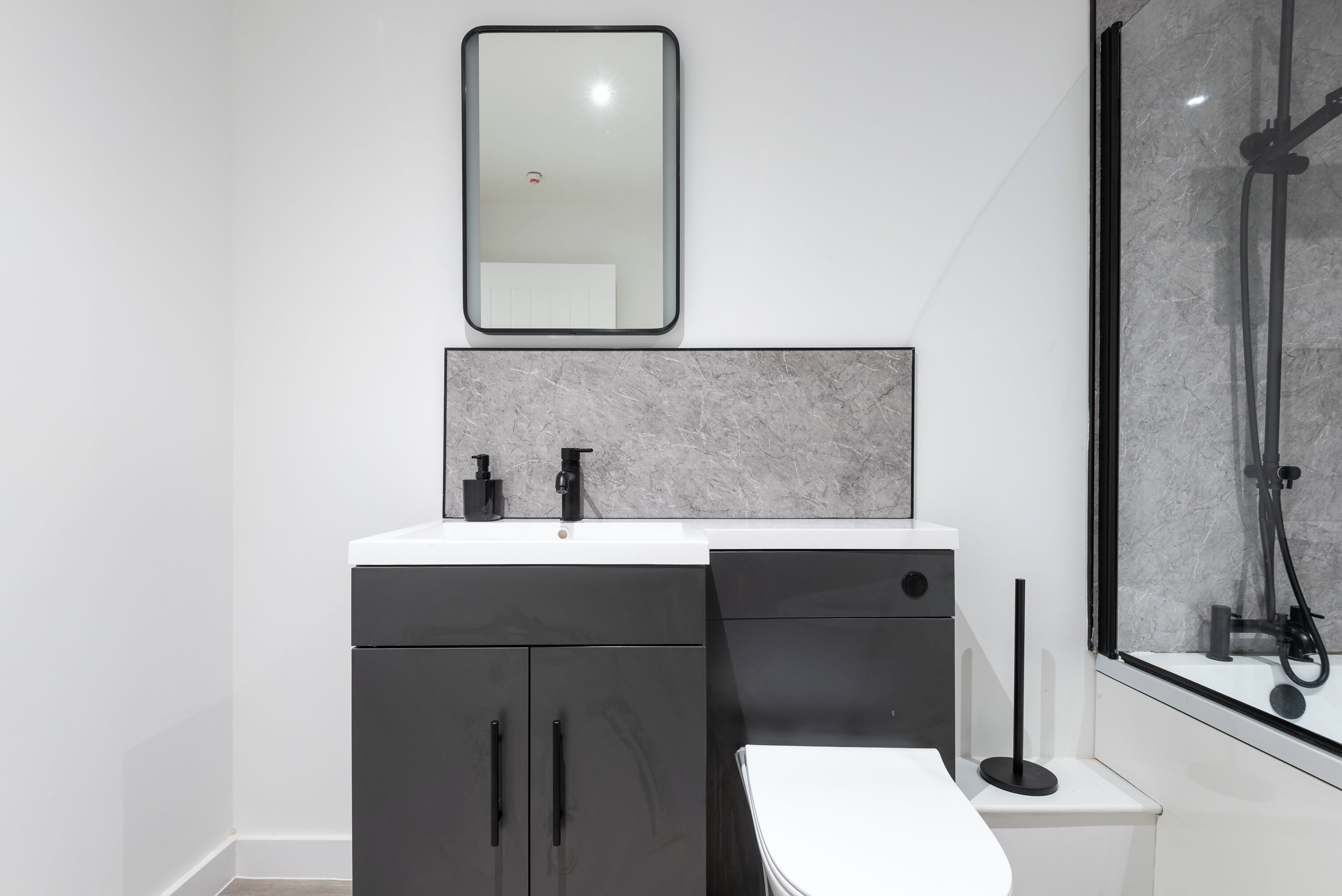 Une salle de bain moderne avec un lavabo blanc, des meubles noirs, un pare-feu en marbre, un miroir et une douche à l’italienne.