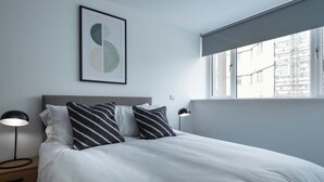 Ein ordentlich bezogenes Bett mit weißer Bettwäsche und gestreiften Kissen, ein Nachttisch mit Lampe, ein gerahmtes abstraktes Kunstwerk an der Wand und ein Fenster mit Blick auf Gebäude.