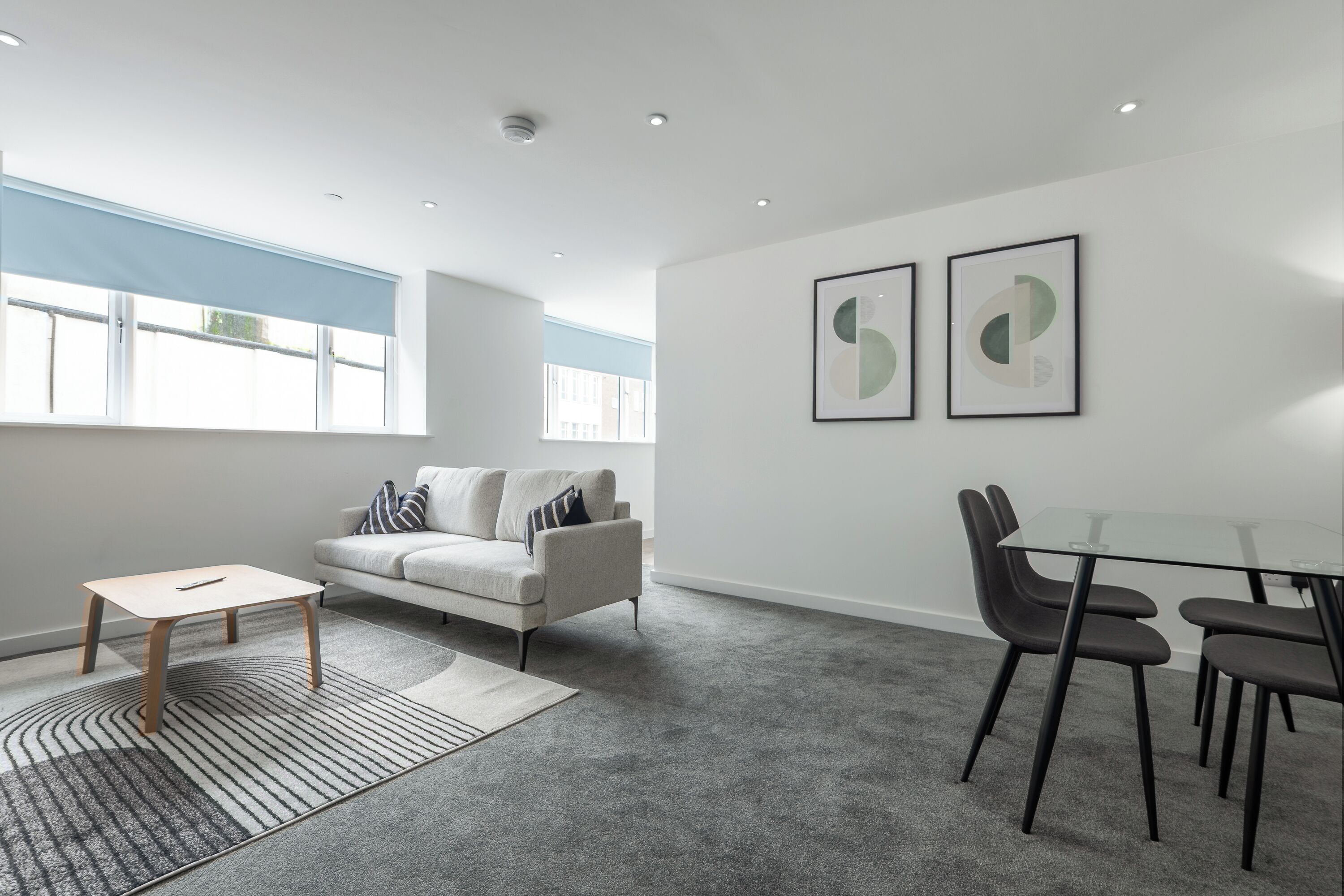 A modern living room with a sofa, a glass table, and chairs, a patterned rug, and two framed abstract artworks on the wall.
