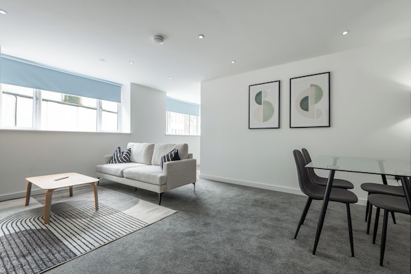 Un salon moderne avec un canapé, une table en verre et des chaises, un tapis à motifs et deux œuvres d’art abstraites encadrées accrochées au mur.