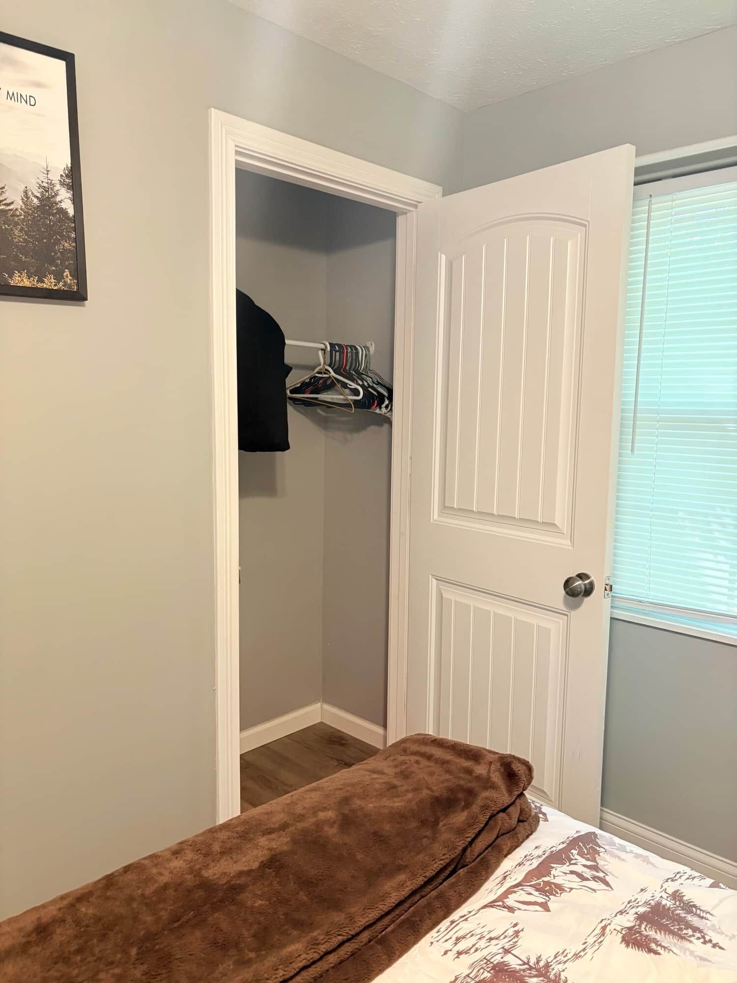 A bedroom with a bed, a closet with hanging clothes, and a door with a window.