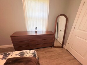 A bedroom with a wooden dresser, a bed with a patterned bedspread, a window with sheer curtains, and a door with a mirror. - Nature's Nook I Cozy 2BR I 15m to RRG I Wi-Fi (Stanton)