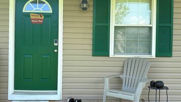 A green door with a welcome sign, a window with green shutters, a white chair, and a small table with a kettle and a pot.