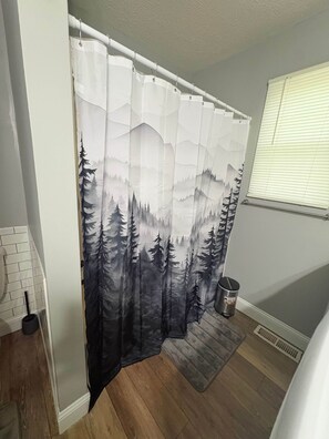A bathroom with a shower curtain featuring a forest scene, a white toilet, a small trash can, and a radiator. - Nature's Nook I Cozy 2BR I 15m to RRG I Wi-Fi (Stanton)