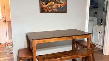 Une table à manger en bois, avec des bancs, dans une pièce aux murs de couleur claire, et on aperçoit un coin cuisine à l’arrière-plan.