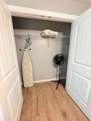 A compact room with a wooden floor, a hanging rack with folded clothes, an ironing board, and a standing fan. - Nature's Nook I Cozy 2BR I 15m to RRG I Wi-Fi (Stanton)